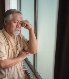 An elderly man in a hospital robe leaning against a window and dealing with the consequences of elder abuse in California.