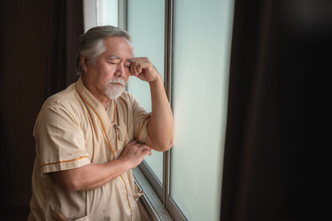An elderly man in a hospital robe leaning against a window and dealing with the consequences of elder abuse in California.