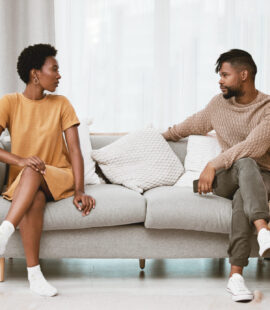 Two siblings contesting a trust, sitting on opposite ends of a couch, one in a dress and the other in a sweater and jeans.